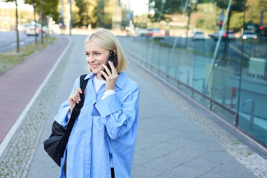 Cheerful young modern woman walking on street and answers phone call, talking on mobile, carries black backpack.