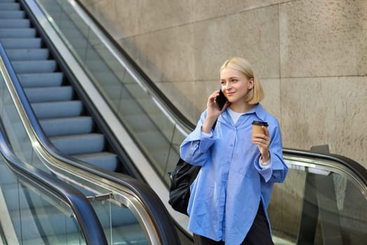 Lifestyle portrait of smiling young woman walking in city, standing on escalator, drinking coffee and chatting on mobile phone.