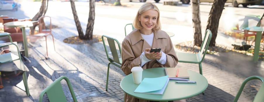 Image of young beautiful girl, student studying, doing homework in cafe, using mobile phone, holding smartphone, smiling.