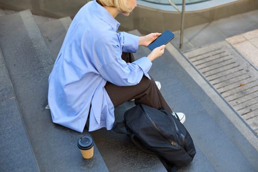 Cropped shot of woman sitting on stairs outside, drinking coffee and looking in smartphone, has backpack near legs.