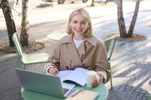 Portrait of beautiful blond woman, wearing wireless headphones, using laptop, studying in outdoor coffee shop, making notes, working on project.