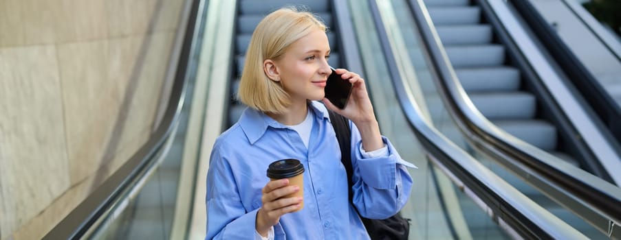 Portrait of stylish young woman with cup of coffee, standing near escalator in city centre, answer phone call, using smartphone, talking to someone.