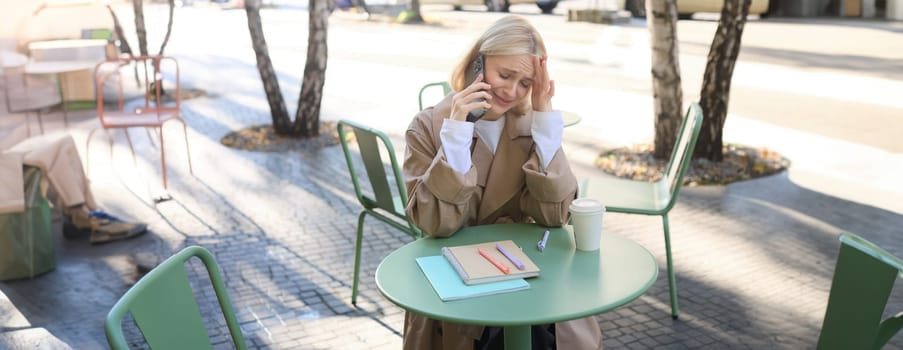 Image of young stressed, upset woman, talking on mobile phone with worried face expression, has difficult conversation over the telephone, sitting in outdoor cafe.