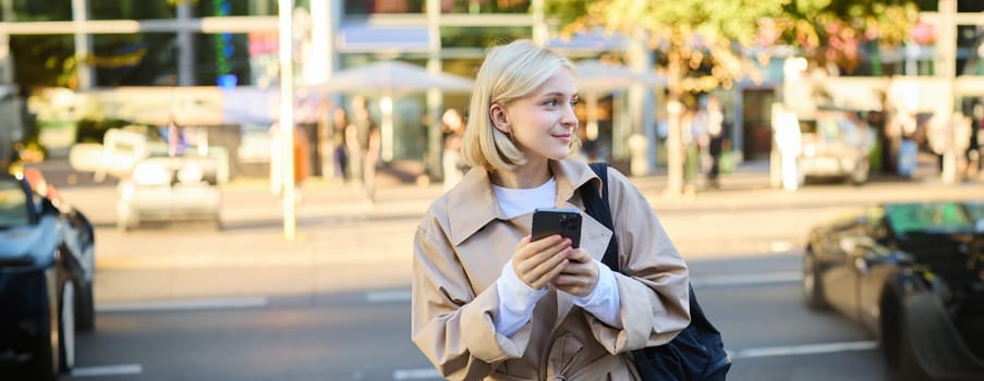 Lifestyle portrait of young business woman, student with backpack, waiting for bus on street, using mobile phone, looking aside and smiling, posing with smartphone.