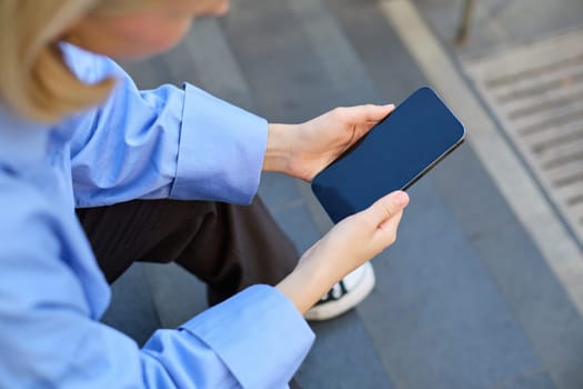 Shot from behind, young woman sitting on stairs on street, holding mobile phone in both hands, screen is dark and blank.