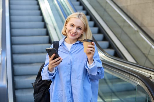 Image of beautiful female model, student standing near escalator, raising cup of takeaway coffee, smiling and looking happy, has mobile phone in hand and backpack on shoulder.