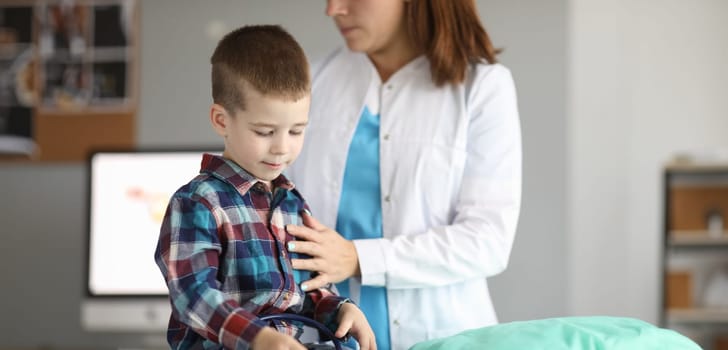 Portrait of child sitting in modern clinic office and looking down in gloomy mood. Female pediatrician standing near patient and examining boy. Medicine and healthcare concept
