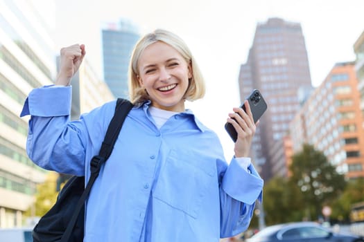 Confident, happy young woman celebrating, raising hand up and looking satisfied, using smartphone, triumphing while standing on empty street. Lifestyle and emotions concept