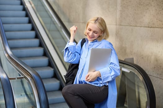 Excited young blond woman with backpack and laptop, says yes, winning, celebrating victory, achieve goal, posing near escalator. Lifestyle concept