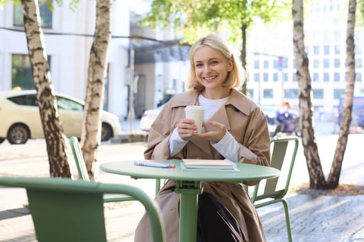 Urban lifestyle concept. Young beautiful woman sitting in outdoor cafe, contemplating city life, drinking coffee and smiling on street.