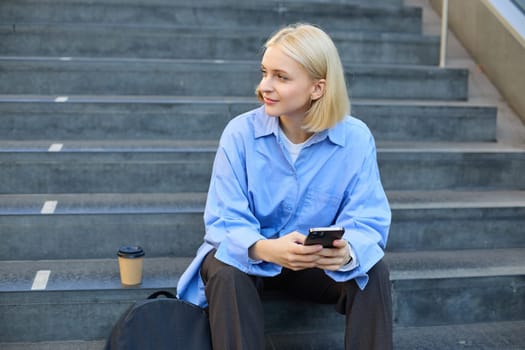 Lifestyle portrait of young urban female model, student drinks cup of takeaway coffee, sitting on stairs outdoors, using mobile phone, smiling and looking happy while taking a break.