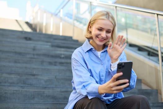 Cute smiling young woman, sitting on stairs with mobile phone, waving hand and saying hello during video chat, connects to online meeting.