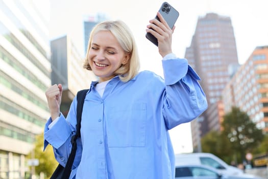 Portrait of happy, enthusiastic young woman, holding backpack and mobile phone, standing on street and rejoicing, triumphing, making fist pump gestures, dancing from happiness and joy.