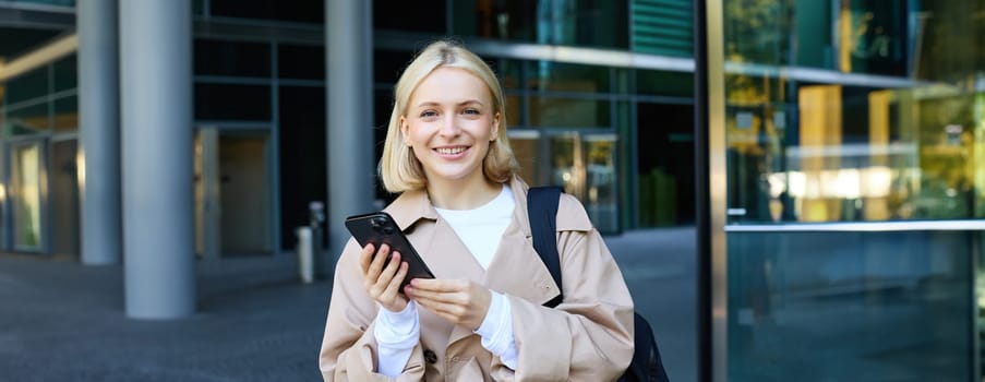 Lifestyle portrait of young beautiful woman, waiting for someone on street, holding smartphone, using mobile phone, ordering taxi in application.