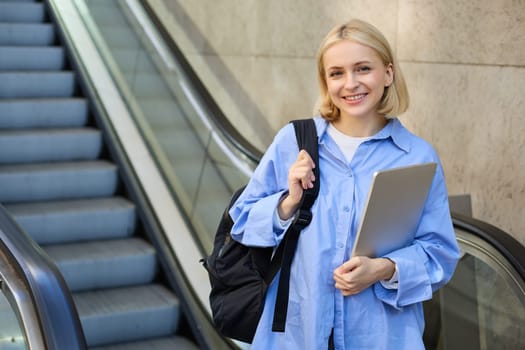 Concept of education and people. Young woman with backpack, carries laptop in hand, standing near escalator, going to tube, on her way to work on university.