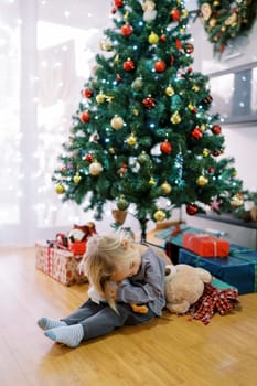 Little girl hugging a toy duck while sitting on the floor near the Christmas tree with gifts. High quality photo