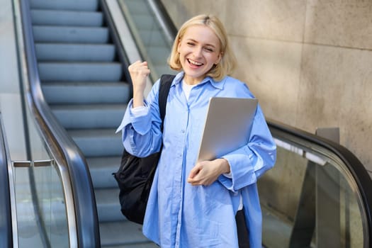 Portrait of enthusiastic young woman, makes fist pump, celebrating or triumphing, holding laptop and backpack, standing near escalator.