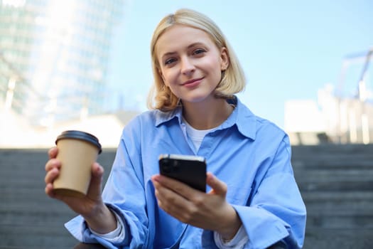 Close up portrait of young smiling woman with cup of coffee, drinking and sitting on stairs in city, holding smartphone in hand.