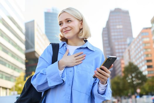 Image of young smiling woman, young professional, standing with backpack and mobile phone, holding hand on chest, looking aside with pleased, happy face expression.