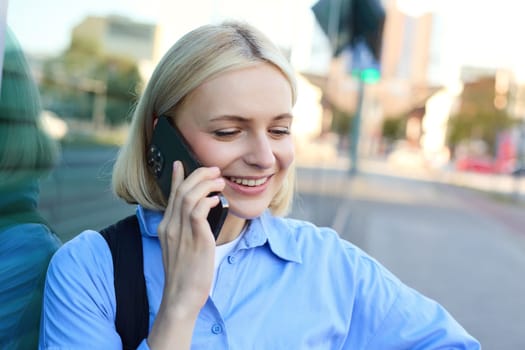 Close up portrait of blond smiling woman, answers a phone call, talking on telephone, standing on street and chatting, looking carefree and happy.