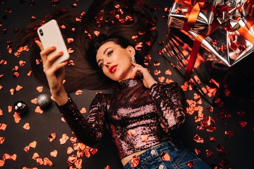 Top view of a girl lying in shiny clothes on the floor in confetti in the form of hearts and taking a selfie.