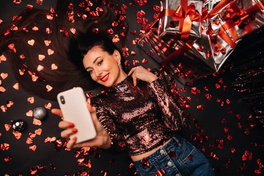 Top view of a girl lying in shiny clothes on the floor in confetti in the form of hearts and taking a selfie.