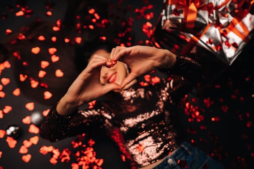 Top view of a girl lying in shiny clothes on the floor in confetti in the form of hearts and showing her heart with her hands.