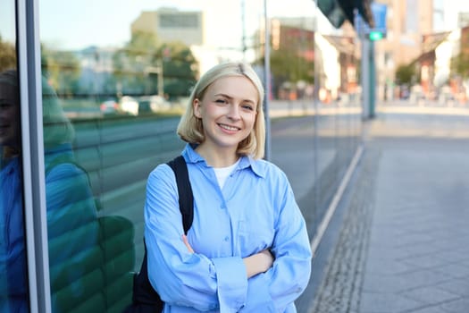 Lifestyle portrait of young smiling woman, student or office employee, standing on street in blue shirt, cross arms on chest and looking confident at camera.