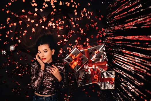 Top view of a girl lying in shiny clothes on the floor in confetti in the form of hearts and gifts.