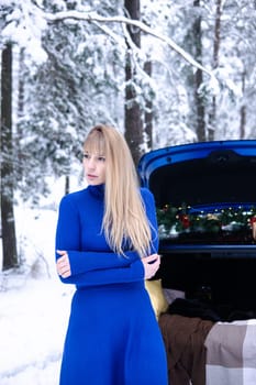 Woman in winter snowy forest in blue dress next to blue car decorated with Christmas decor. Christmas and winter holidays concept