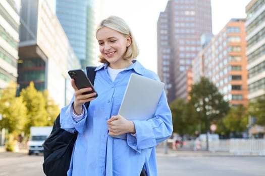 Image of young stylish, modern blond woman in blue shirt, holding laptop and backpack, using smartphone, smiling, walking along the street, posing in city centre.