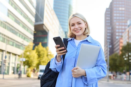 Cheerful smiling blond woman with mobile phone, posing with laptop in city centre, concept of new horizons and education opportunities. Lifestyle portrait