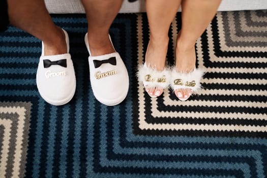 Male and female feet in slippers stand on a striped carpet. Caption: Groom. Bride. Cropped, faceless. High quality photo