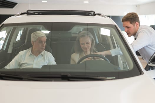 A gray-haired Caucasian woman is sitting behind the wheel of a car. Car dealership salesman consulting mature married couple