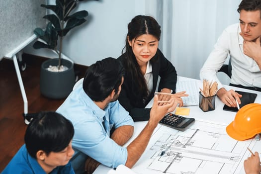 Diverse group of civil engineer and client working together on architectural project, reviewing construction plan and building blueprint at meeting table. Prudent