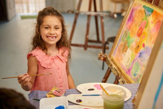 Caucasian cute elementary age child, little girl artist smiles with a beautiful toothy smile, looking at the camera, holding paintbrush and drawing on canvas while learning fine art in creative studio