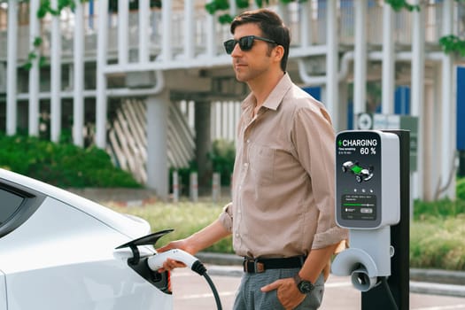 Young man put EV charger to recharge electric car's battery from charging station in city commercial parking lot. Rechargeable EV car for sustainable environmental friendly urban travel. Expedient