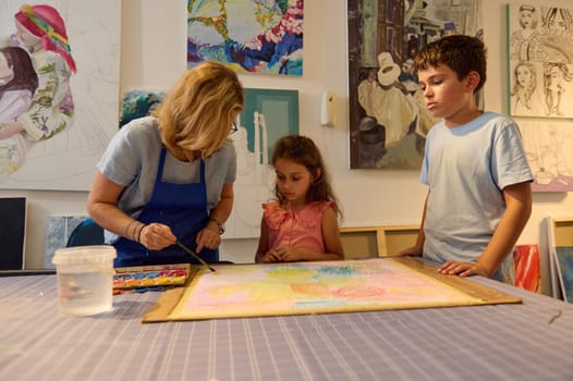 In cozy creative studio group of elementary age children attend art class led by inspired artist woman, teaching preschoolers to paint pictures on canvas. Kids development, educational courses, hobby