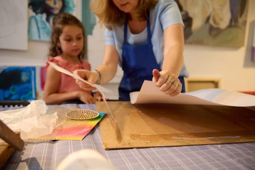 Details on white cardboard paper sheet on the desk against blurred background of a paint teacher and her little student drawing working in creative art workshop. Kids, creativity and education concept