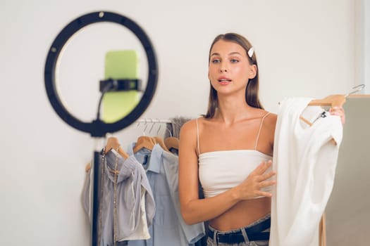 Young woman making fashion video content for social media. Blogger smiles to camera using selfie stick and light ring while making persuasive online clothing sell to audience or followers. Unveiling