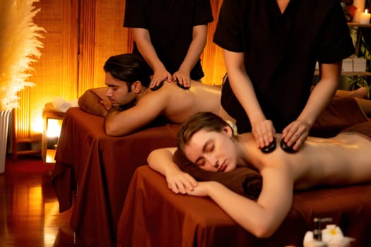 Hot stone massage at spa salon in luxury resort with warm candle light, blissful couple customer enjoying spa basalt stone massage glide over body with soothing warmth. Quiescent