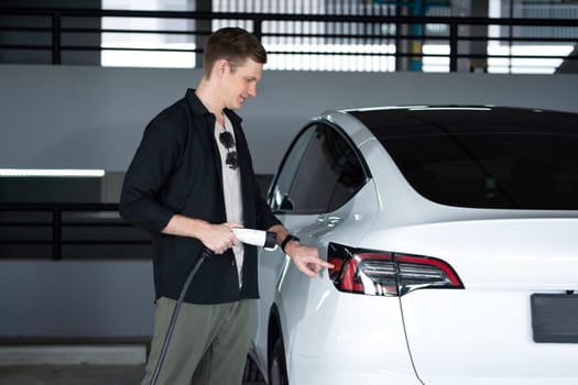 Young man travel with EV electric car to shopping center parking lot charging in downtown city showing urban sustainability lifestyle by green clean rechargeable energy of electric vehicle innards