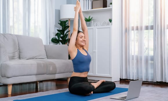 Asian woman in sportswear doing yoga exercise on fitness mat as her home workout training routine. Healthy body care lifestyle woman watching online yoga video on laptop. Vigorous