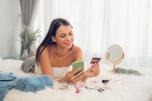 Young woman lying on the bed using online payment app and digital wallet on smartphone to pay with credit card. E commerce shopping and modern purchasing via mobile internet. Unveiling