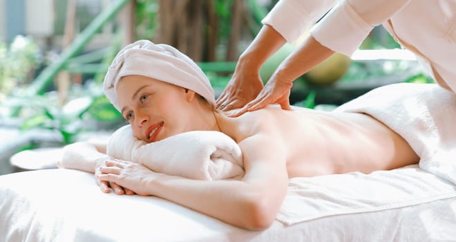 Beautiful young woman received a back massage on a spa bed from professional masseuse. Attractive female relaxes deeply by skilled hands of the massage therapist. Surrounded with nature. Tranquility.