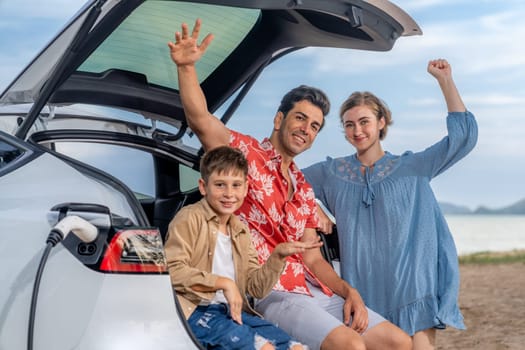 Family vacation trip traveling by the beach with electric car, lovely family sit on the trunk, charging EV car battery with green and sustainable energy. Family travel and eco-friendly car. Perpetual