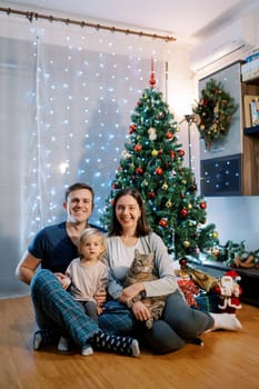 Smiling mom and dad with a cat and a little daughter on their knees sit on the floor near the Christmas tree. High quality photo