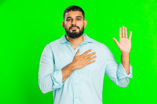 I swear to be honest. Sincere responsible Indian man raising hand to take oath, promising to be honest, to tell truth, keeping hand on chest, justice. Guy isolated on green chroma key background