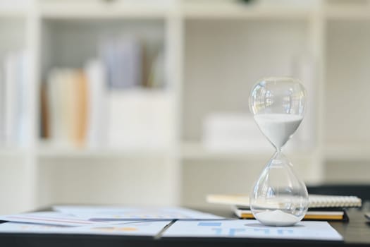 Hourglass on wooden office table with financial documents. Time management concept.