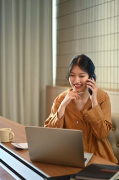 Happy Asian businesswoman having phone conversation and using laptop at modern office.